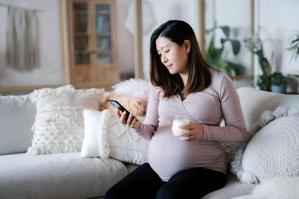 A pregnant woman sitting on a couch at home, booking her pregnancy safe facial appointment on her phone.