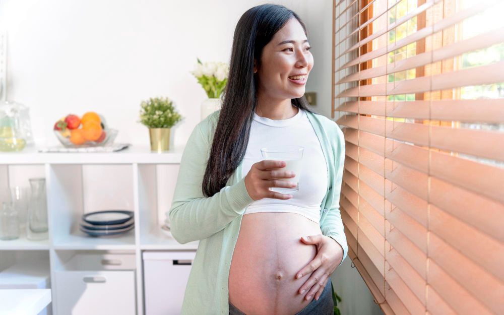 A pregnant woman holding a glass of water in front of a window while enjoying a pregnancy-safe facial treatment.